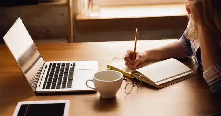 Blogger writing a new blog post on a laptop while taking notes in a notebook.